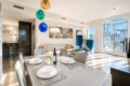 Bright open-plan living and dining area with a glossy table set for six, hanging colorful glass pendants, and a city-view balcony—sunlit and modern.