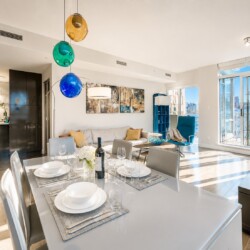 Bright open-plan living and dining area with a glossy table set for six, hanging colorful glass pendants, and a city-view balcony—sunlit and modern.