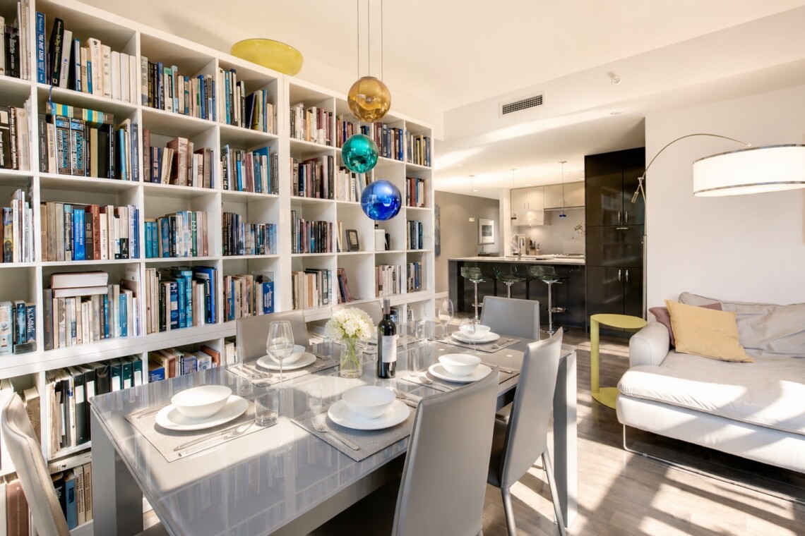 Open-concept living and dining space with a floor-to-ceiling white bookshelf wall, a glass dining table set for six, and colorful pendant lights above the table.