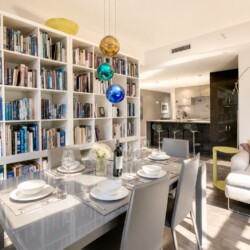 Open-concept living and dining space with a floor-to-ceiling white bookshelf wall, a glass dining table set for six, and colorful pendant lights above the table.