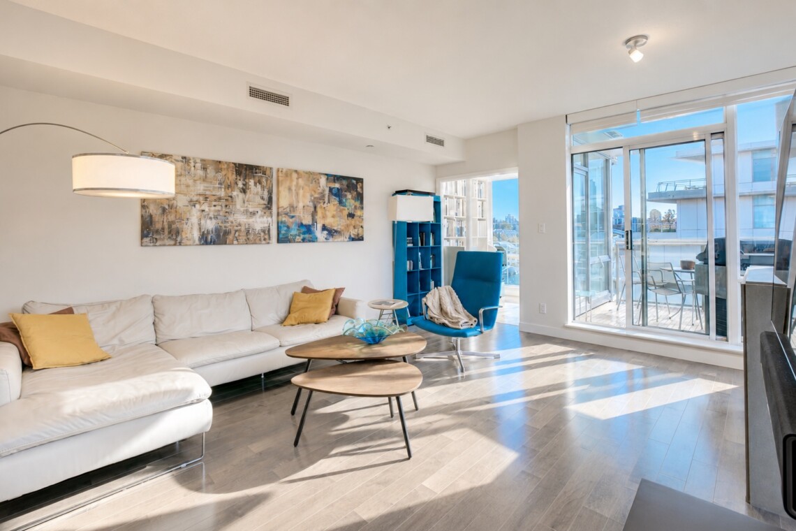 Bright living room with a white sectional sofa and yellow throw pillows, abstract wall art, and a blue accent chair by a two-tier wooden coffee table. A glass door leads to a sunlit balcony.