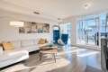 Bright living room with a white sectional sofa and yellow throw pillows, abstract wall art, and a blue accent chair by a two-tier wooden coffee table. A glass door leads to a sunlit balcony.