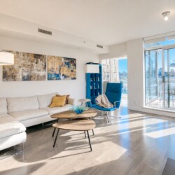Bright living room with a white sectional sofa and yellow throw pillows, abstract wall art, and a blue accent chair by a two-tier wooden coffee table. A glass door leads to a sunlit balcony.
