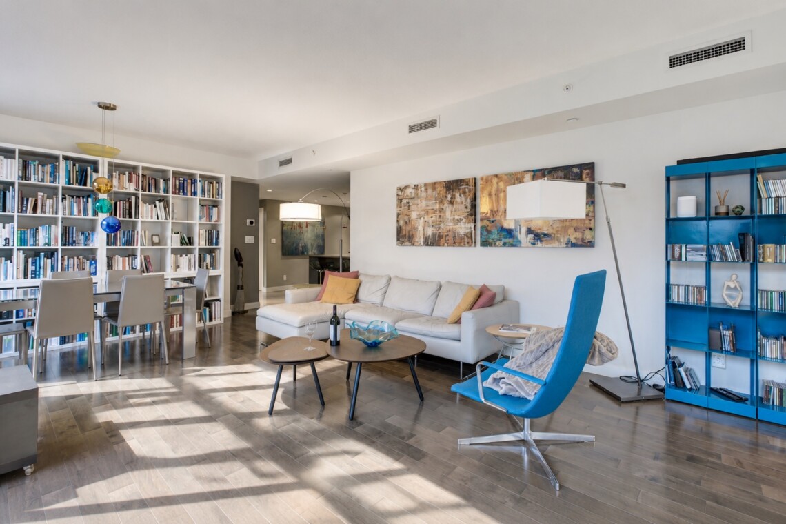 Bright, modern living space with a white sofa, blue lounge chair, and tall white bookcases full of books on the left.