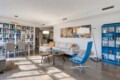 Bright, modern living space with a white sofa, blue lounge chair, and tall white bookcases full of books on the left.