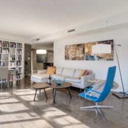 Bright, modern living space with a white sofa, blue lounge chair, and tall white bookcases full of books on the left.