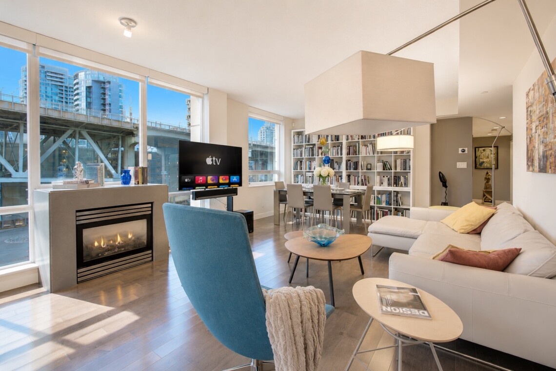 Bright open living room with a fireplace, TV, blue accent chair, white sectional sofa, and a tall bookshelf by large windows overlooking the city.