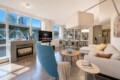 Bright open living room with a fireplace, TV, blue accent chair, white sectional sofa, and a tall bookshelf by large windows overlooking the city.