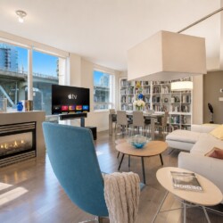 Bright open living room with a fireplace, TV, blue accent chair, white sectional sofa, and a tall bookshelf by large windows overlooking the city.