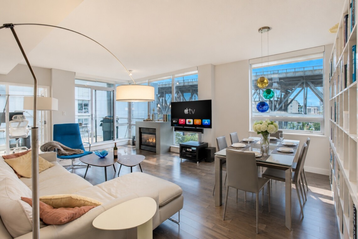 Bright open-plan living room with a beige sectional, blue lounge chair, fireplace, and a wall-mounted TV near a glass balcony door. It also shows a dining area with a table and chairs.