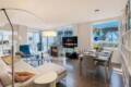 Bright open-plan living room with a beige sectional, blue lounge chair, fireplace, and a wall-mounted TV near a glass balcony door. It also shows a dining area with a table and chairs.
