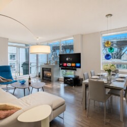 Bright open-plan living room with a beige sectional, blue lounge chair, fireplace, and a wall-mounted TV near a glass balcony door. It also shows a dining area with a table and chairs.