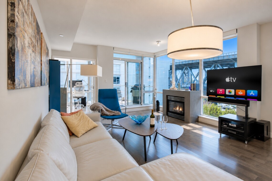 Bright, modern living room with a white sectional, blue accent chair, fireplace, and a large TV by floor-to-ceiling windows nearby balconies/urban view.