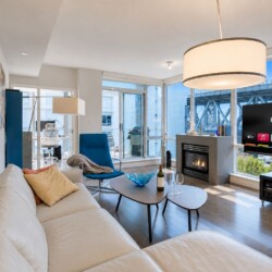 Bright, modern living room with a white sectional, blue accent chair, fireplace, and a large TV by floor-to-ceiling windows nearby balconies/urban view.
