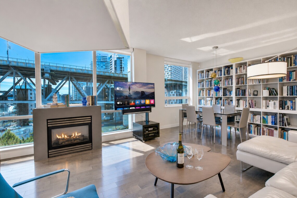 Bright open-concept living room with a gas fireplace, large windows overlooking a cityscape, and a wall of bookshelves; a TV on a stand and a glass dining table nearby.