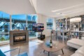 Bright open-concept living room with a gas fireplace, large windows overlooking a cityscape, and a wall of bookshelves; a TV on a stand and a glass dining table nearby.