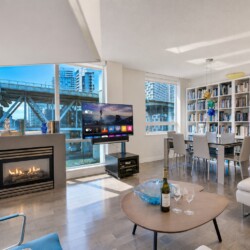 Bright open-concept living room with a gas fireplace, large windows overlooking a cityscape, and a wall of bookshelves; a TV on a stand and a glass dining table nearby.