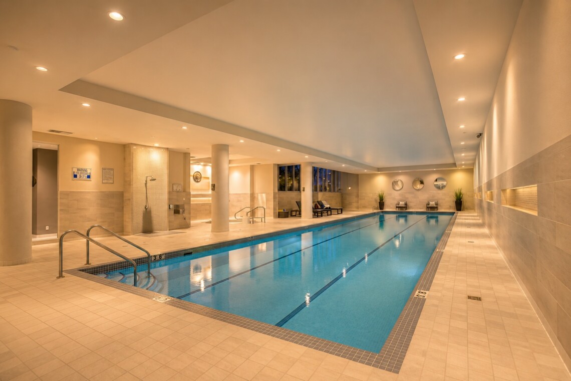 Indoor pool area with beige tile floors and walls, soft overhead lighting, and lounge chairs along the far wall; a calm blue rectangular pool in the center. Depth markers run along the pool edge.