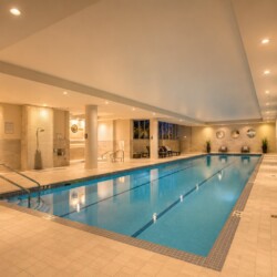 Indoor pool area with beige tile floors and walls, soft overhead lighting, and lounge chairs along the far wall; a calm blue rectangular pool in the center. Depth markers run along the pool edge.
