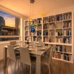 Sunset-lit dining room with a glass table set for six, a white bookshelf wall, and colorful hanging glass orbs by the ceiling window.