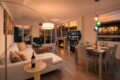 Open-plan living and dining area at sunset: white sofa with pastel cushions, round coffee table with snacks and wine, fireplace, TV, and a glass dining table set for six near large windows.