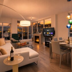 Open-plan living and dining area at sunset: white sofa with pastel cushions, round coffee table with snacks and wine, fireplace, TV, and a glass dining table set for six near large windows.