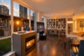 Open-plan living room at sunset with a gas fireplace, large windows, and a wall of bookshelves facing a TV on a stand.
