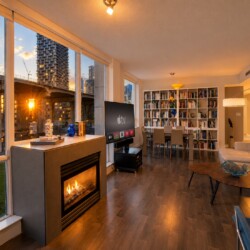 Open-plan living room at sunset with a gas fireplace, large windows, and a wall of bookshelves facing a TV on a stand.