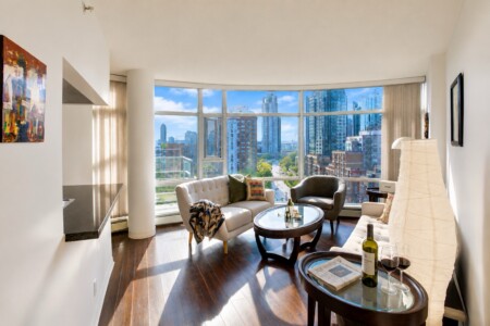 Bright, modern living room with curved windows and a city skyline view, white tufted sofa, gray chairs, and a round glass coffee table.