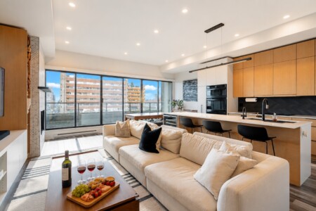 Bright open-concept living room and kitchen with a beige sectional, wine and snacks on a wooden tray, and large city-view windows.
