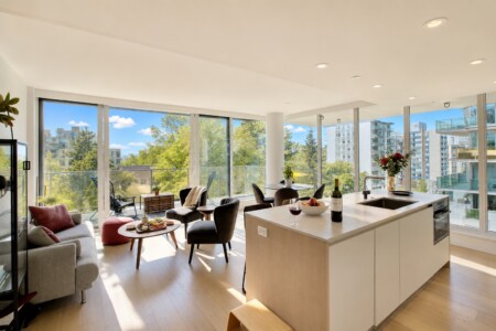 Bright open-plan living area with floor-to-ceiling windows, a balcony, and a modern kitchen island with wine and fruit.