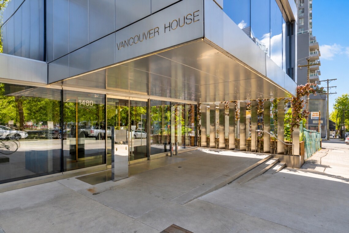 Front entrance of Vancouver House with glass doors, a metallic canopy, and reflective windows at 1480, showing trees and street reflections.