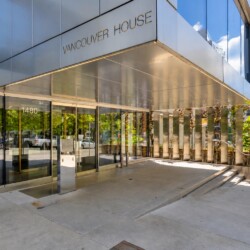 Front entrance of Vancouver House with glass doors, a metallic canopy, and reflective windows at 1480, showing trees and street reflections.