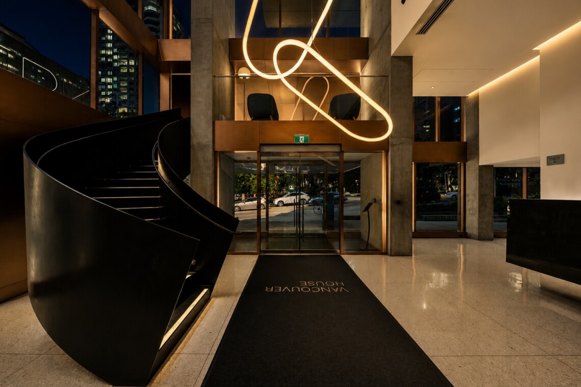 Modern hotel lobby with a curved black staircase, glass entrance, and a glowing loop sculpture overhead.