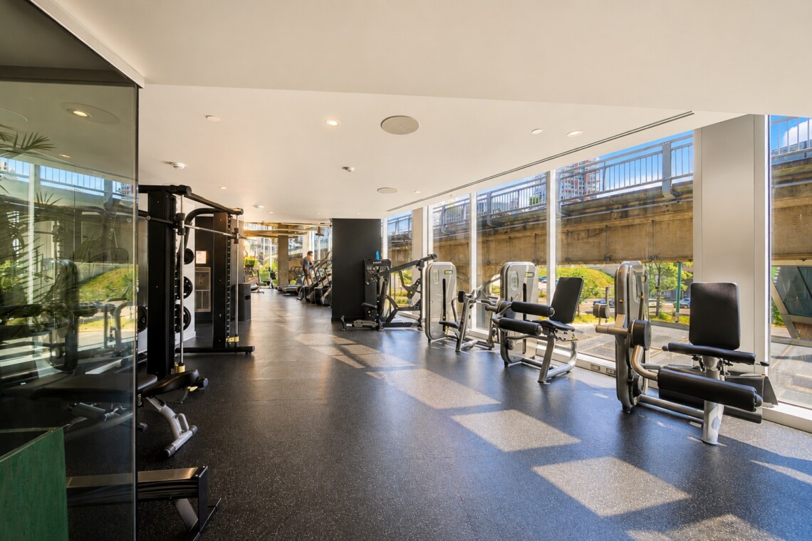 Bright gym interior with weight machines and cardio equipment along large windows overlooking an outdoor railing area.