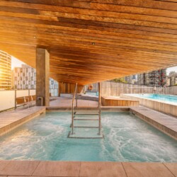 Rooftop pool under a warm wooden canopy, with a metal ladder and city high-rises in the background at sunset.