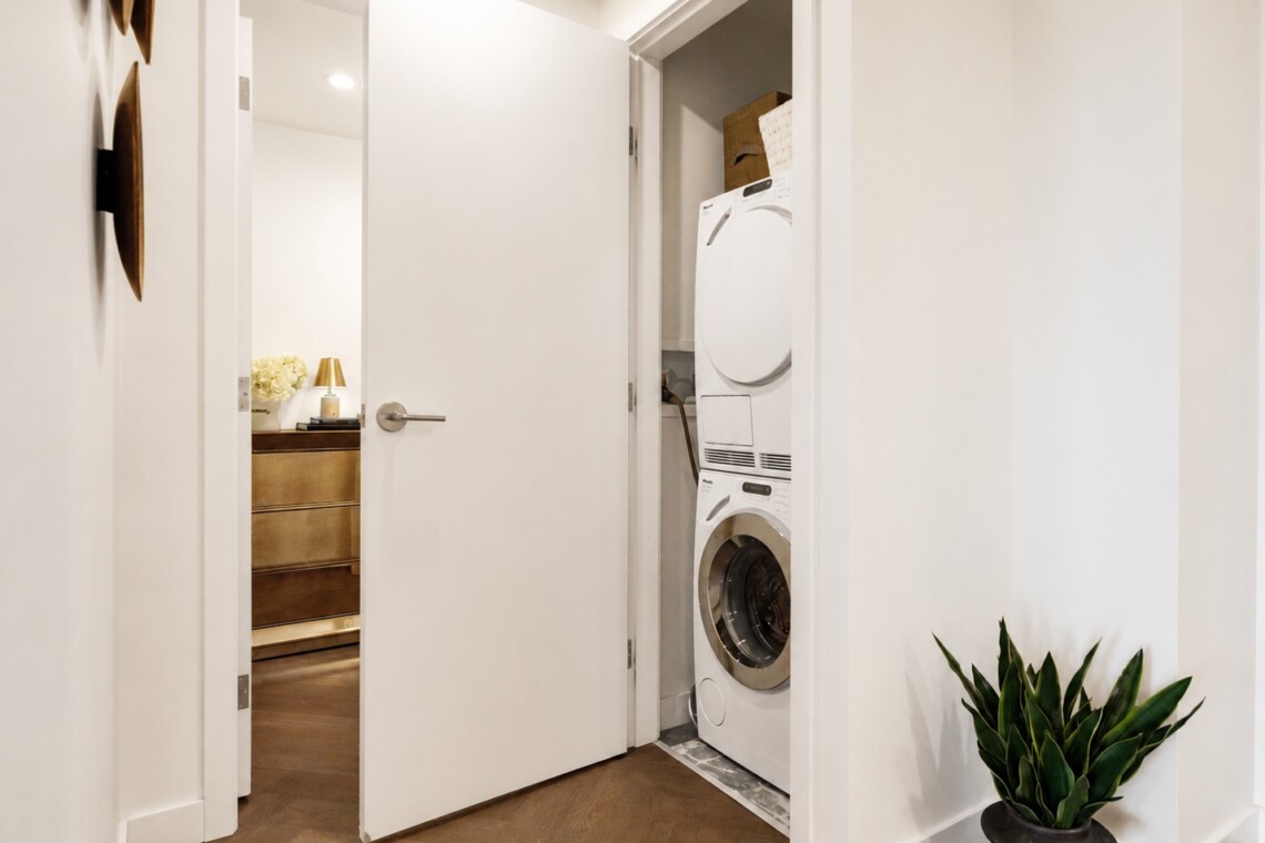 Open door reveals a laundry closet with a stacked washer and dryer in white cabinetry and a potted plant nearby in the hallway.