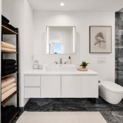 Modern bathroom with a white floating vanity, backlit mirror, and glass-walled shower on dark marble floors and walls, plus a wall-mounted toilet.
