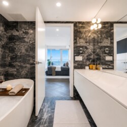 Modern bathroom with black marble walls, white vanity, and a bathtub; a wooden tray holds wine, a candle, and a rolled towel.