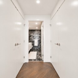 White-paneled hallway with doors on both sides, opening to a marble-tiled bathroom at the end; wooden floor in foreground.