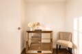 Gold mirrored dresser with flowers and a small lamp in a bright corner, beige upholstered chair to the right.