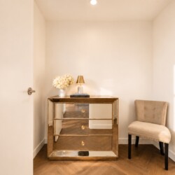 Gold mirrored dresser with flowers and a small lamp in a bright corner, beige upholstered chair to the right.