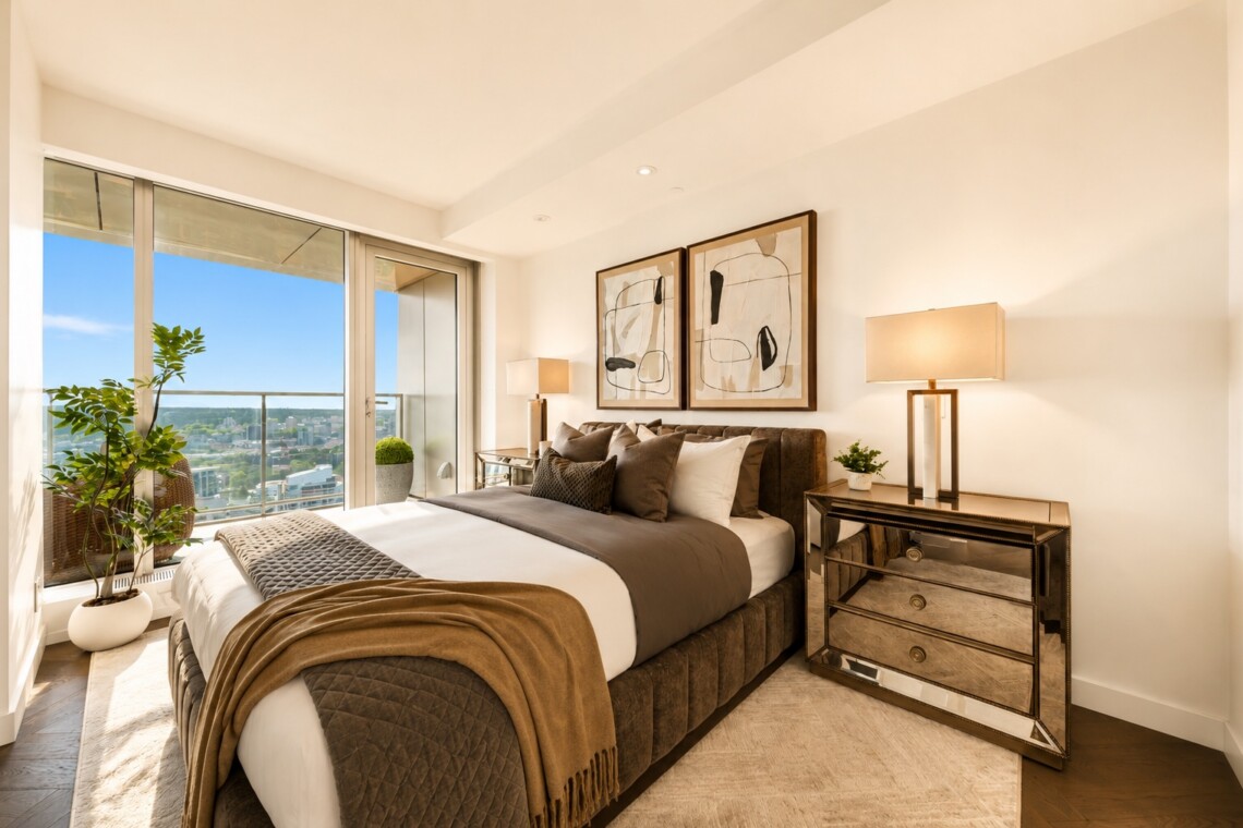 Bright modern bedroom with a large window and city view; upholstered bed with brown/gray bedding, abstract art above, and a mirrored dresser with lamps.