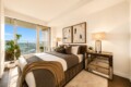 Bright modern bedroom with a large window and city view; upholstered bed with brown/gray bedding, abstract art above, and a mirrored dresser with lamps.