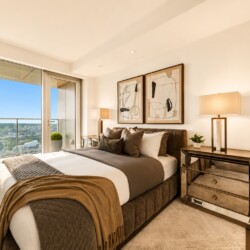 Bright modern bedroom with a large window and city view; upholstered bed with brown/gray bedding, abstract art above, and a mirrored dresser with lamps.