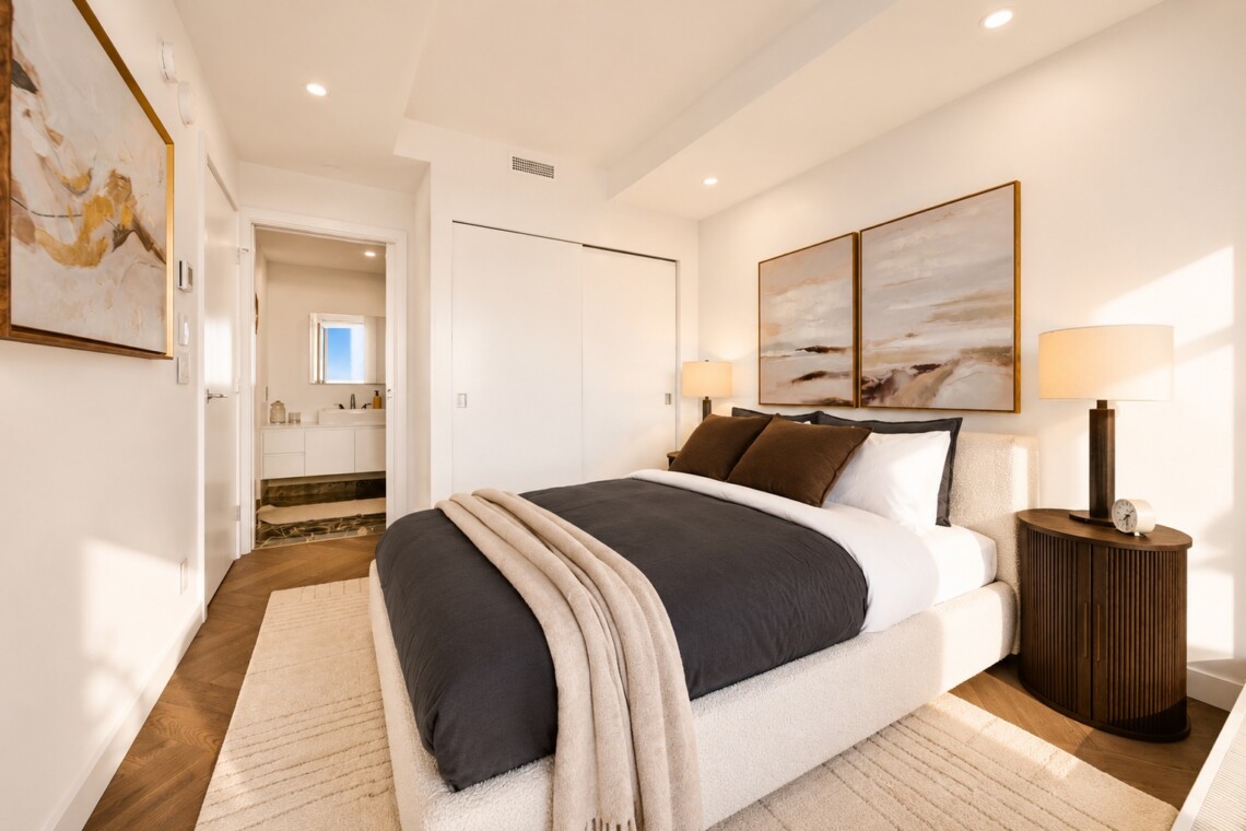 Contemporary bedroom with a plush upholstered bed, dark gray comforter, beige throw, two framed landscape paintings, and a round wooden nightstand with lamp; doorway to a bath visible in the background.
