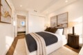 Contemporary bedroom with a plush upholstered bed, dark gray comforter, beige throw, two framed landscape paintings, and a round wooden nightstand with lamp; doorway to a bath visible in the background.
