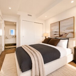 Contemporary bedroom with a plush upholstered bed, dark gray comforter, beige throw, two framed landscape paintings, and a round wooden nightstand with lamp; doorway to a bath visible in the background.