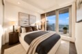 Cozy modern bedroom with a low platform bed, dark bedding, beige throw, and matching nightstands, opening to a balcony with a city view behind glass doors.