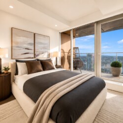 Cozy modern bedroom with a low platform bed, dark bedding, beige throw, and matching nightstands, opening to a balcony with a city view behind glass doors.